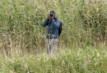 Featured Bird Watcher: Rudy Chodankar Photographer in a blue jacket using a DSLR camera and binoculars in a tall grass field.