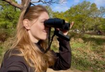 Featured Bird Watcher: Becca Heywood Smiling blonde woman looking through black binoculars in a sunny green park.