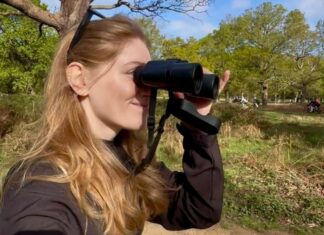 Featured Bird Watcher: Becca Heywood Smiling blonde woman looking through black binoculars in a sunny green park.